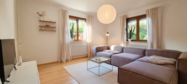 Wohnzimmer mit großem Ecksofa, Glastisch, Teppich und zwei Fenstern